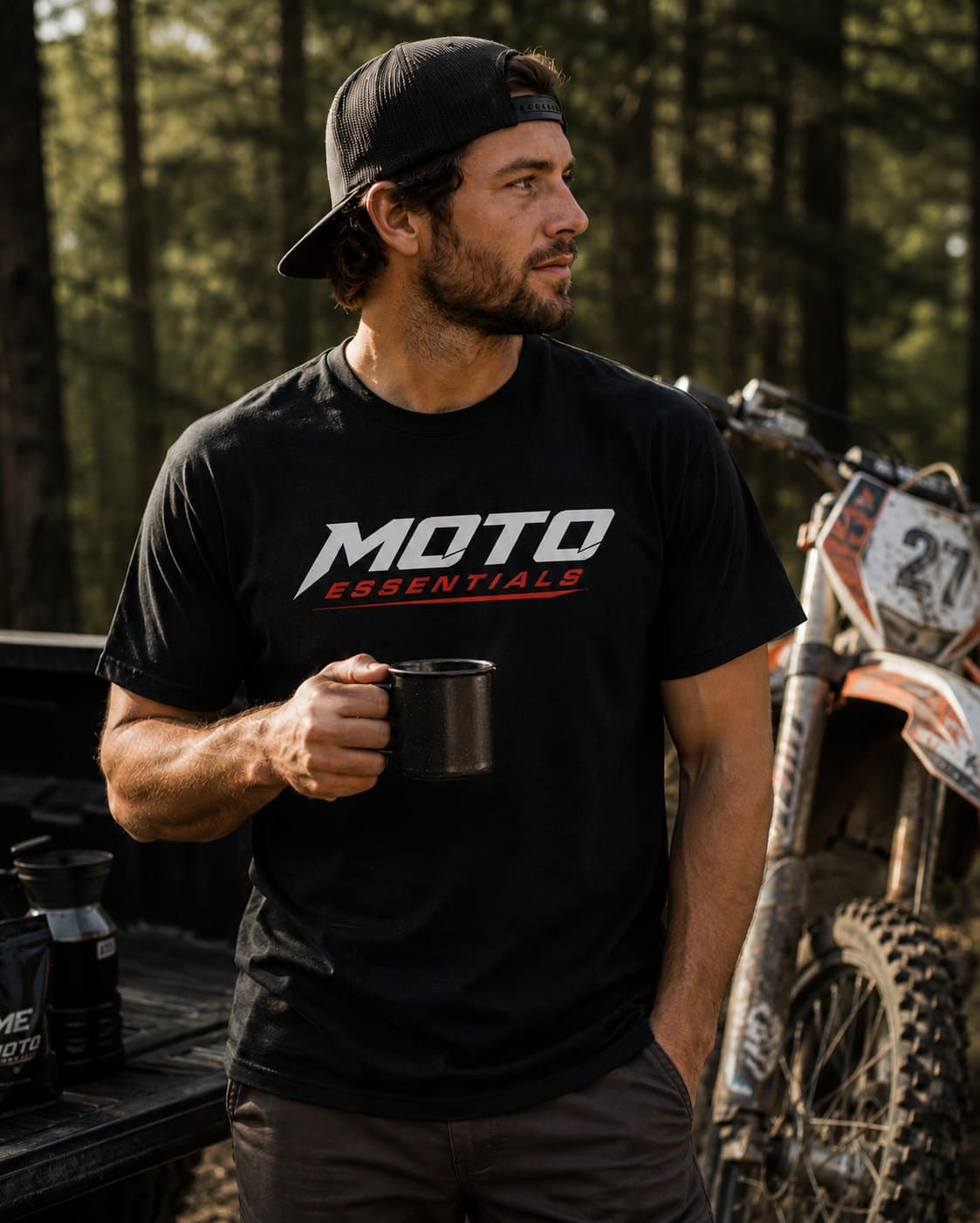 Rider wearing the black Moto Essentials logo tee, holding a camp mug beside a dirt bike in the woods
